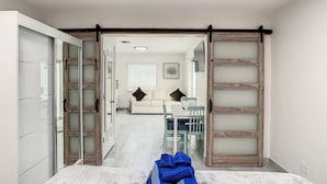 Habitación de hotel con cama, armario, zona de comedor con mesa y sillas, y sala de estar con sofá y televisor.