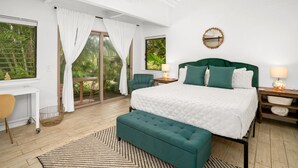 Une chambre avec un grand lit, un bureau et une chaise. Il y a une fenêtre avec des rideaux, un ventilateur de plafond et une vue sur la verdure à l’extérieur.