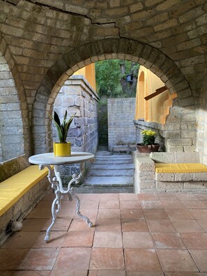An arched stone corridor with a small table and bench, potted plants, and a staircase leading to a higher level.