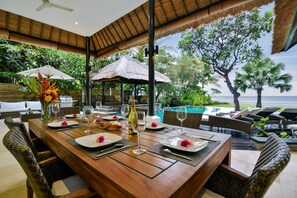 A dining area with a wooden table set for a meal, surrounded by wicker chairs, with a view of a pool and tropical vegetation.