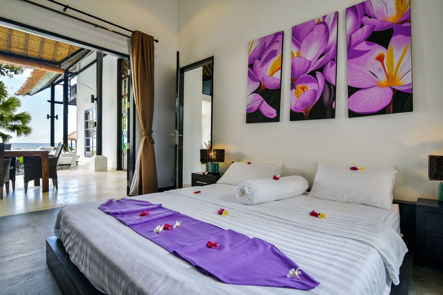 A hotel room with a large bed, a dining area with a view of the sea, and three paintings of flowers on the wall.