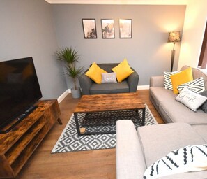 Una sala moderna con un sofá gris, una mesa de centro de madera y un mueble para la televisión. Hay cojines decorativos y una alfombra estampada en el suelo.