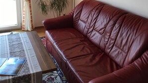 A living room with a red leather sofa, a patterned table, a floor lamp, a potted plant, and a framed floral painting on the wall. - Vacation apartment for up to 4 people, 90 sqm, 2 large bedrooms, balcony (Piding)