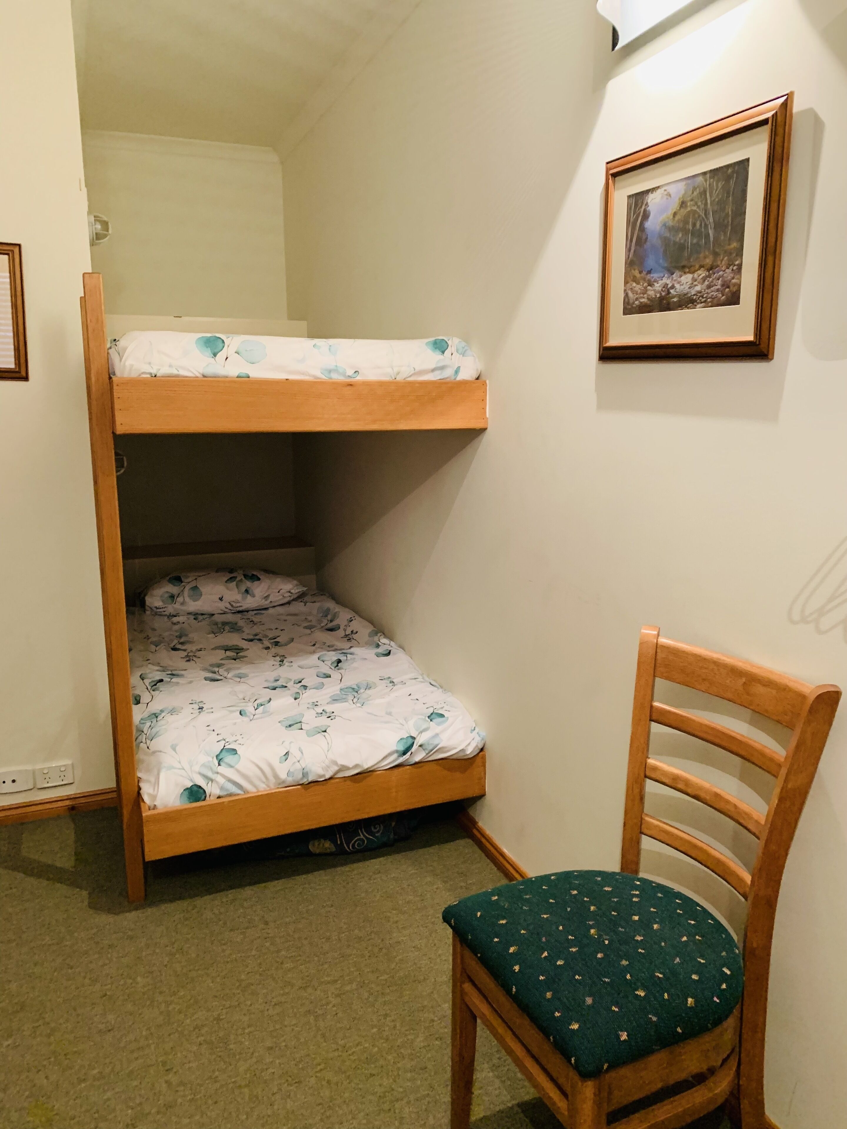A bunk bed with floral bedding, a wooden chair with a green cushion, and a framed picture on the wall.