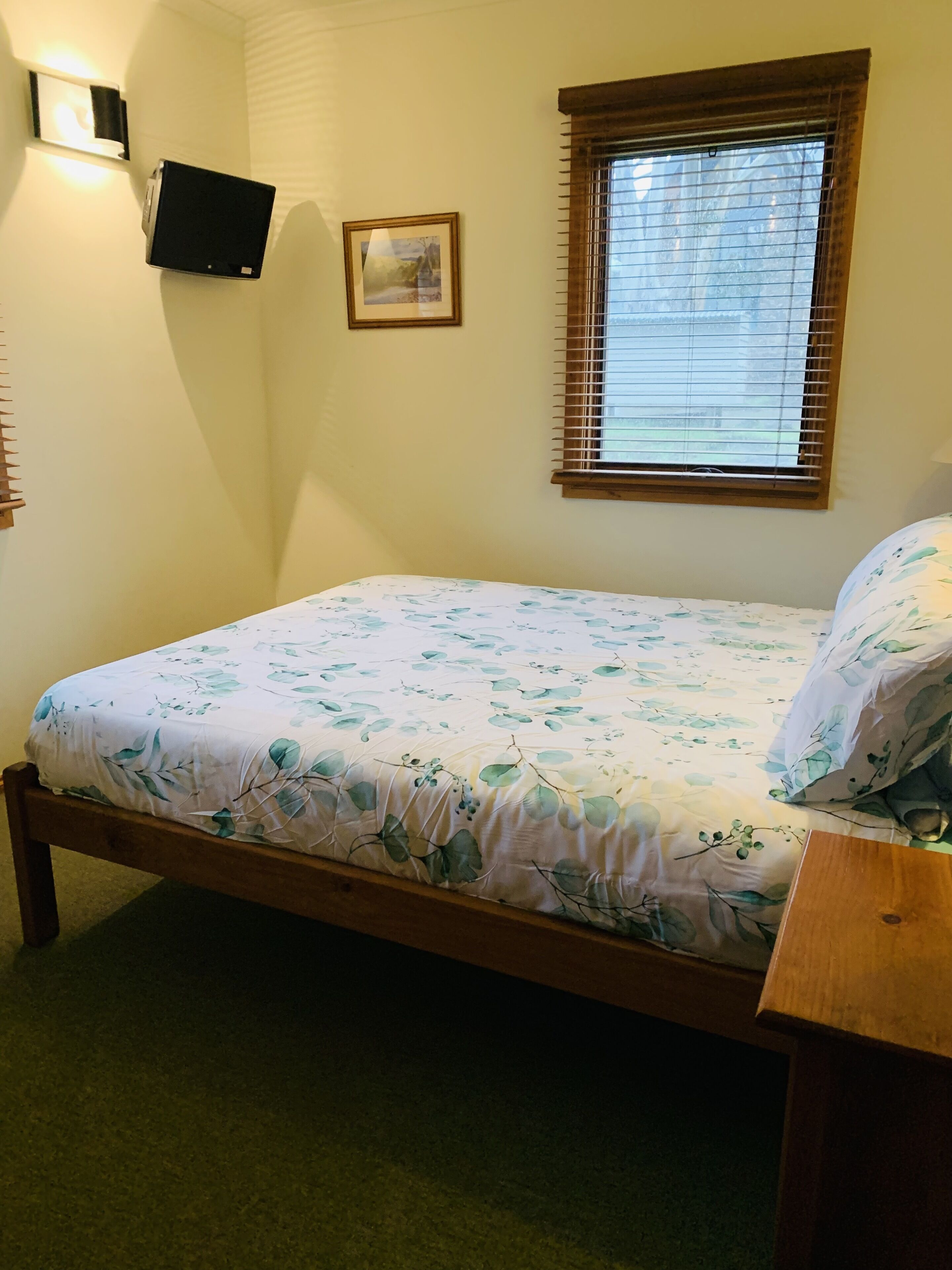 Una camera da letto con un letto, una TV a parete, una finestra con persiane e un quadro appeso al muro.