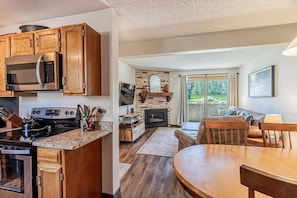 A modern kitchen with a built-in microwave and stove, a dining area with a wooden table, and a living room with a fireplace and a large window.