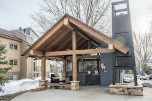 A covered outdoor area with a wooden structure, stone benches, and a grill.