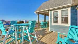 Eine Holzterrasse mit türkisfarbenen Stühlen und Tischen, ein überdachter Bereich mit Geländer und Blick auf den Ozean.