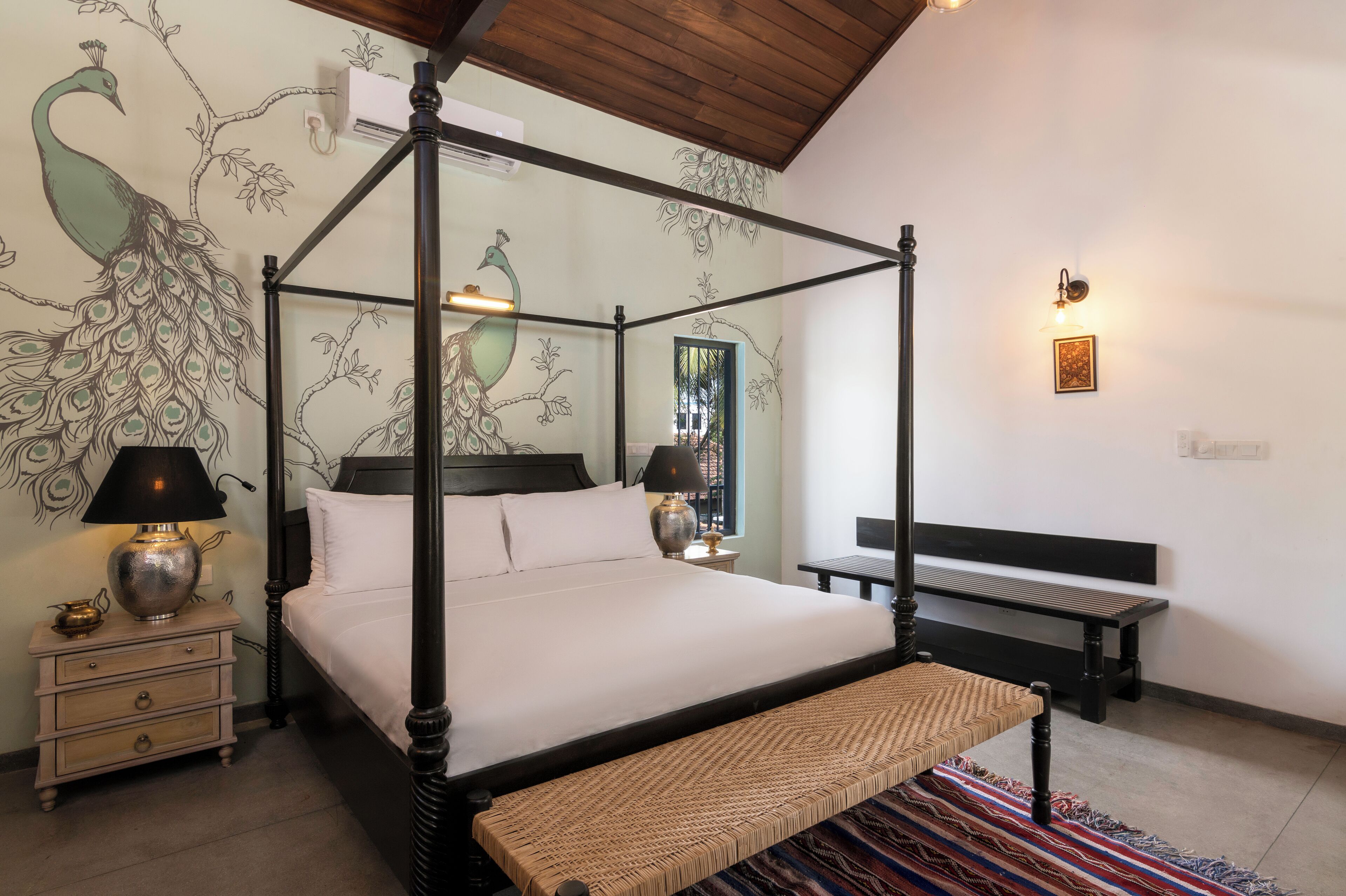 Una cama con dosel, un banco tejido, una mesita de noche con lámpara y un mural en la pared con pavos reales.