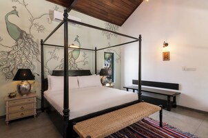 Una cama con dosel, un banco tejido, una mesita de noche con lámpara y un mural en la pared con pavos reales.