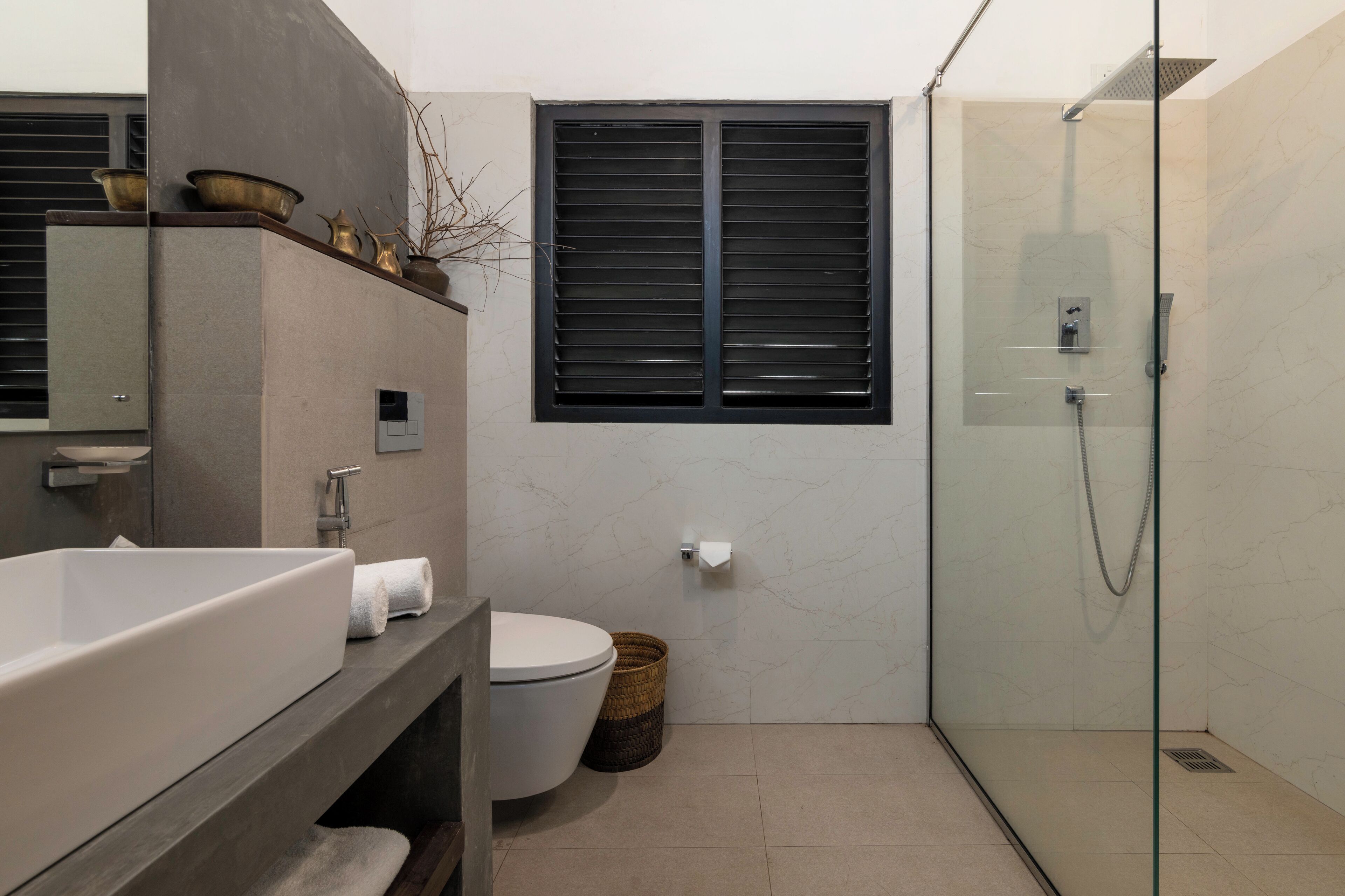 A modern bathroom with a large white sink, a toilet, a glass-enclosed shower, and a window with black blinds.