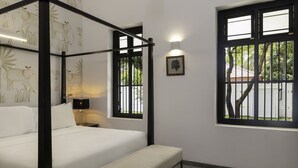 A four-poster bed with a headboard, a bedside table with a lamp, and a window with a view of greenery and a building outside.