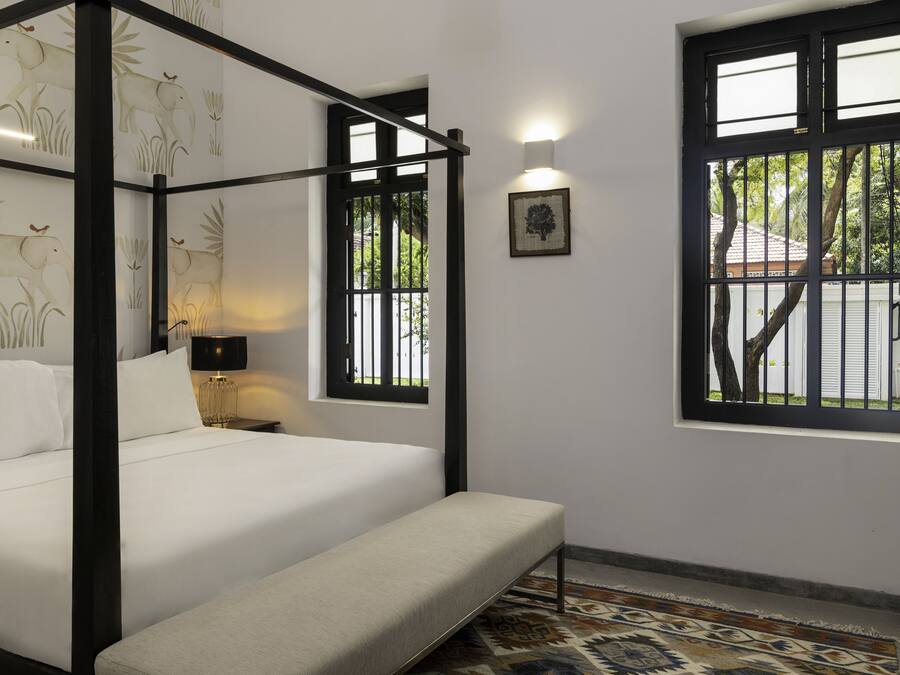 A four-poster bed with a headboard, a bedside table with a lamp, and a window with a view of greenery and a building outside.