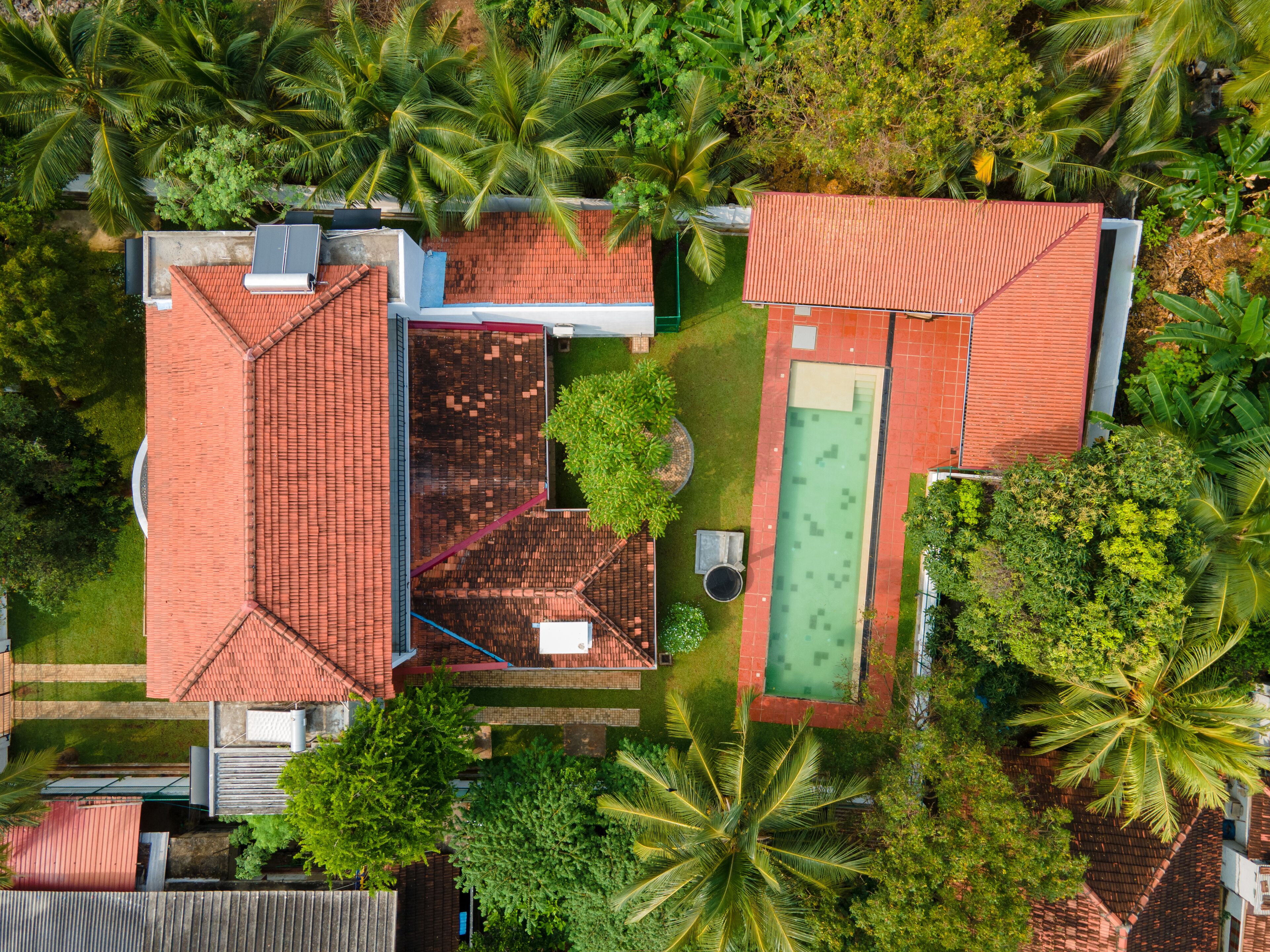 An aerial view of a residential area with two houses, each with a red-tiled roof, surrounded by lush greenery and palm trees.