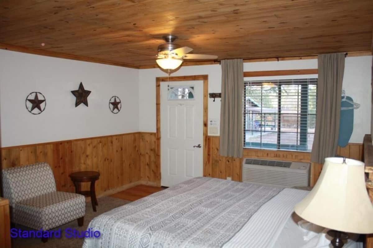 A room with a wooden ceiling, a bed with a patterned bedspread, a chair, a small table, and a window with curtains.