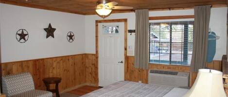 A room with a wooden ceiling, a bed with a patterned bedspread, a chair, a small table, and a window with curtains.