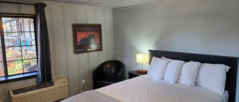 A hotel room with a bed, a chair, a nightstand, a framed picture, and a window with curtains.