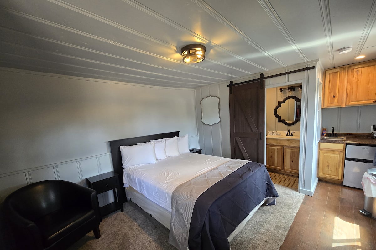 A bedroom with a large bed, a nightstand, a black chair, and a wooden door leading to a kitchen.