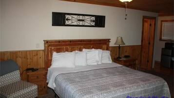 A bedroom with a wooden headboard, a patterned bedspread, a nightstand with a lamp, and a chair.