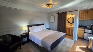 A bedroom with a large bed, a bedside table, a lamp, and a black leather chair. There is a kitchen area visible through an open door. - Lumbermens Village (Lakeside)