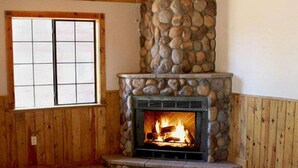 A stone fireplace with a fire, wooden walls, and a window with a grid pattern. - Lumbermens Village (Lakeside)