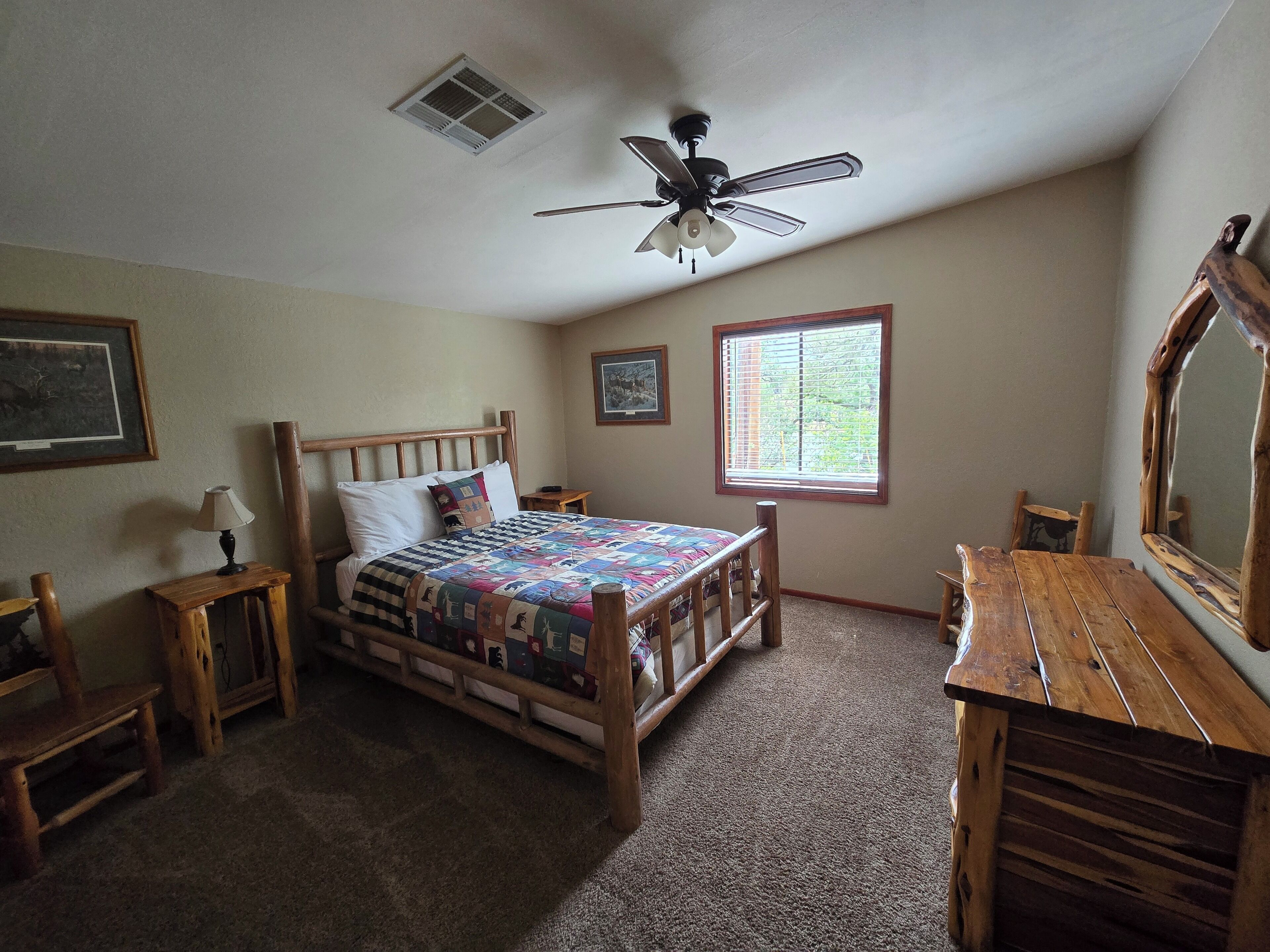 A bedroom with a wooden bed, a ceiling fan, a window with blinds, a wooden dresser, and a mirror.