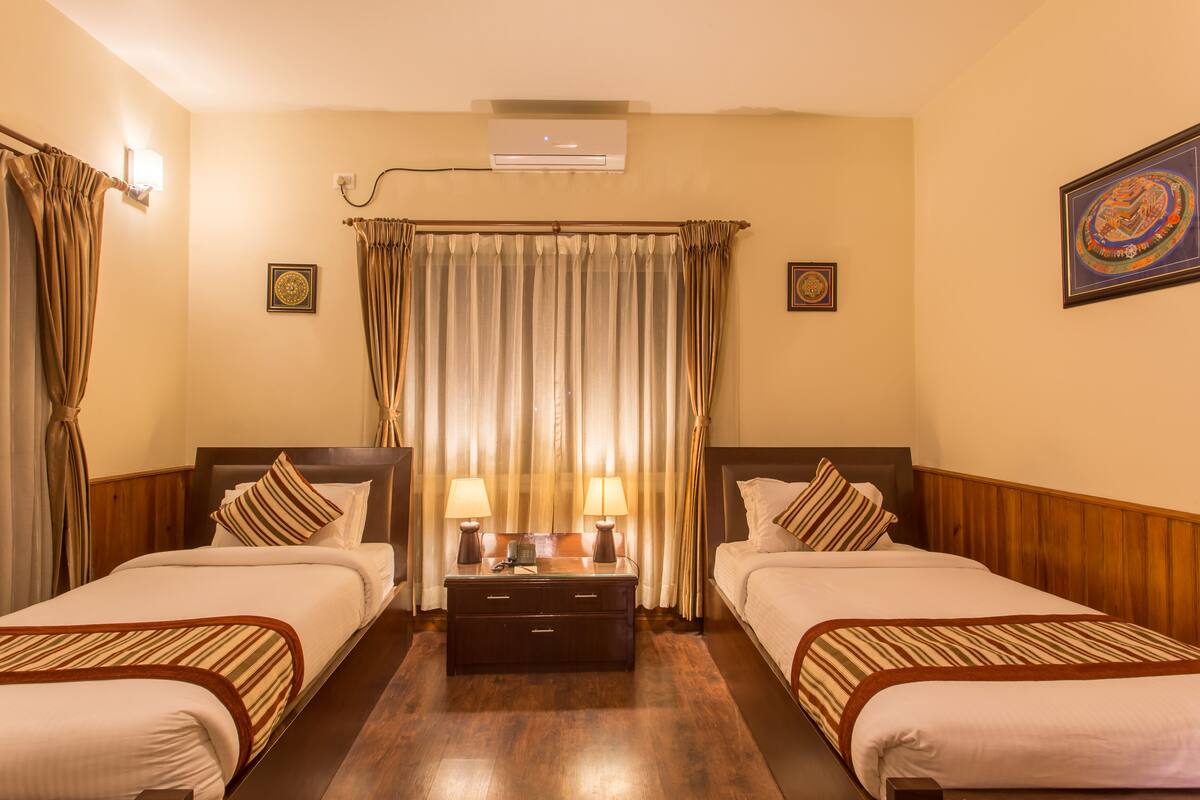 Quarto de hotel com duas camas, mesa de cabeceira, abajur, ar condicionado e cortinas.