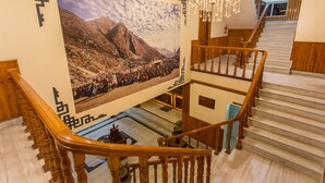Ein mehrgeschossiges Interieur mit hölzernen Treppen, einem Chandelier und einem großen Wandgemälde, das eine gebirgige Landschaft zeigt.