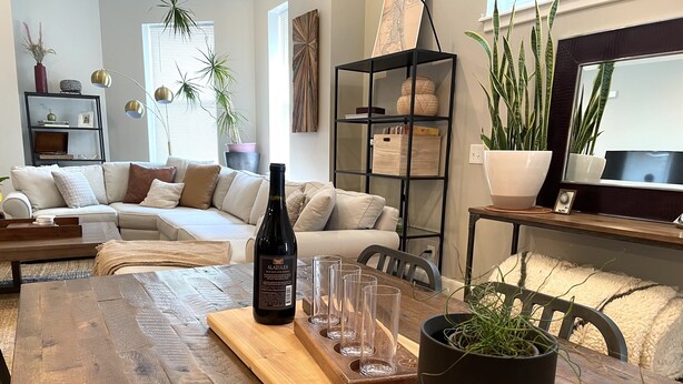 A dining area with a wooden table, a bottle of wine, and glasses. There is a sofa and a bookshelf in the background.