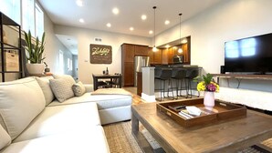 A modern living room with a sofa, a coffee table, and a TV. There is a kitchen area with a refrigerator, microwave, and bar stools.