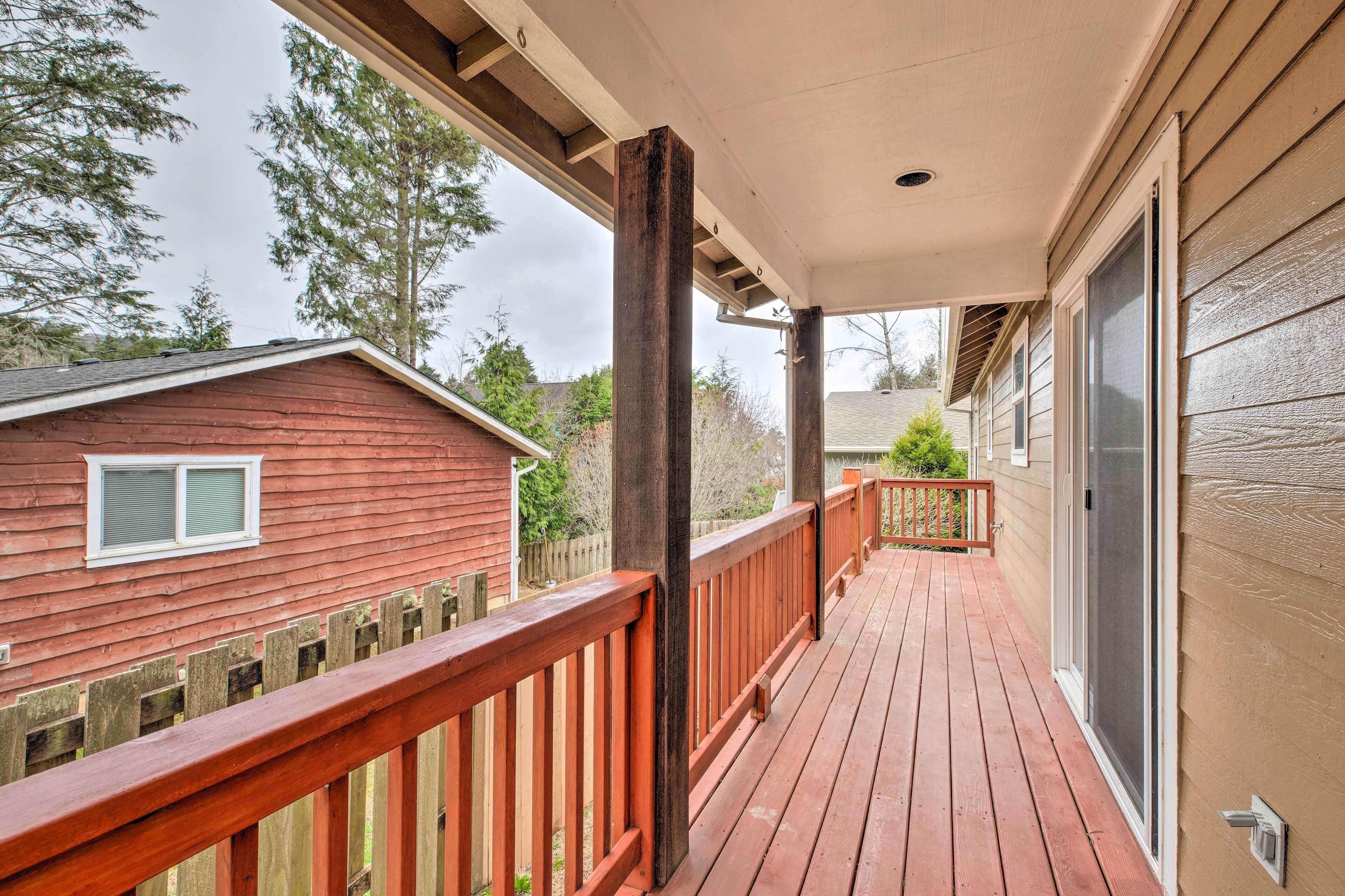 A wooden deck with a railing, a covered porch, and a view of a red building and trees.