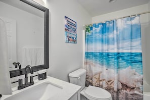 Casa de banho com lavatório branco, espelho, sanita e cortina de chuveiro com paisagem praiana.