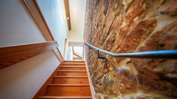 A wooden staircase with a metal handrail, leading up to a room with a stone wall.