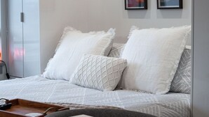 A neatly made bed with white bedding and a decorative pillow, a wooden tray with a book and remote, and two framed artworks on the wall. - Lake Okanagan beach condo w/ private buoy, AC, heated outdoor pool, patio, SUPs (Vernon)