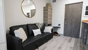 A modern living room with a black sofa, a round mirror, and a wooden cabinet. - Lake Okanagan beach condo w/ private buoy, AC, heated outdoor pool, patio, SUPs (Vernon)