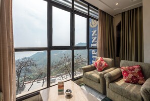 Habitación de hotel con un ventanal grande, dos sillones, una mesita y vistas a un paisaje montañoso.