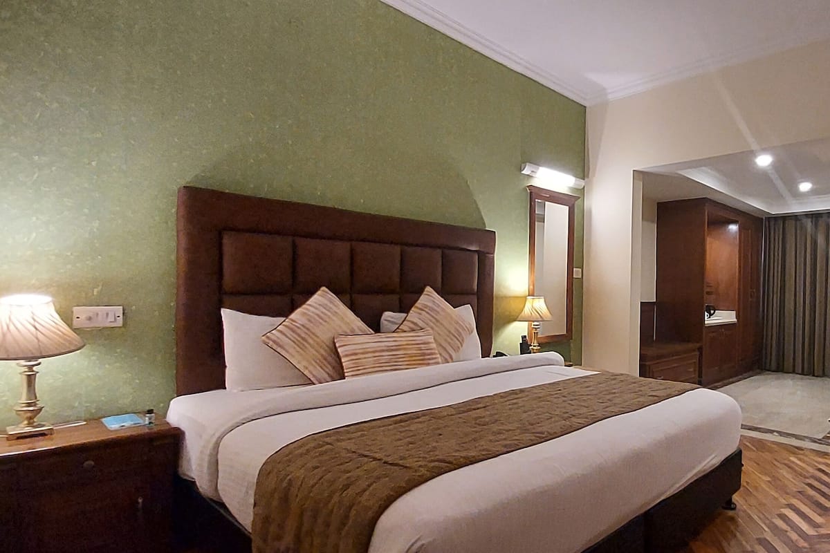 Ein Hotelzimmer mit einem großen Bett, einem Nachttisch mit Lampe, einem Holz-Kopfteil, einem Spiegel, einem Kleiderschrank und einem gerahmten Kunstwerk an der Wand.