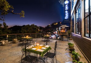 Un espacio al aire libre con mesas dispuestas para dos personas, rodeado de plantas en macetas y vistas a la ciudad de noche.