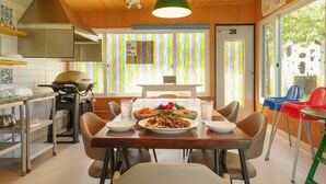 Un comedor con una mesa de madera dispuesta para cuatro personas, rodeada de sillas. Se ve una parrilla al fondo y cortinas de colores a través de la ventana.