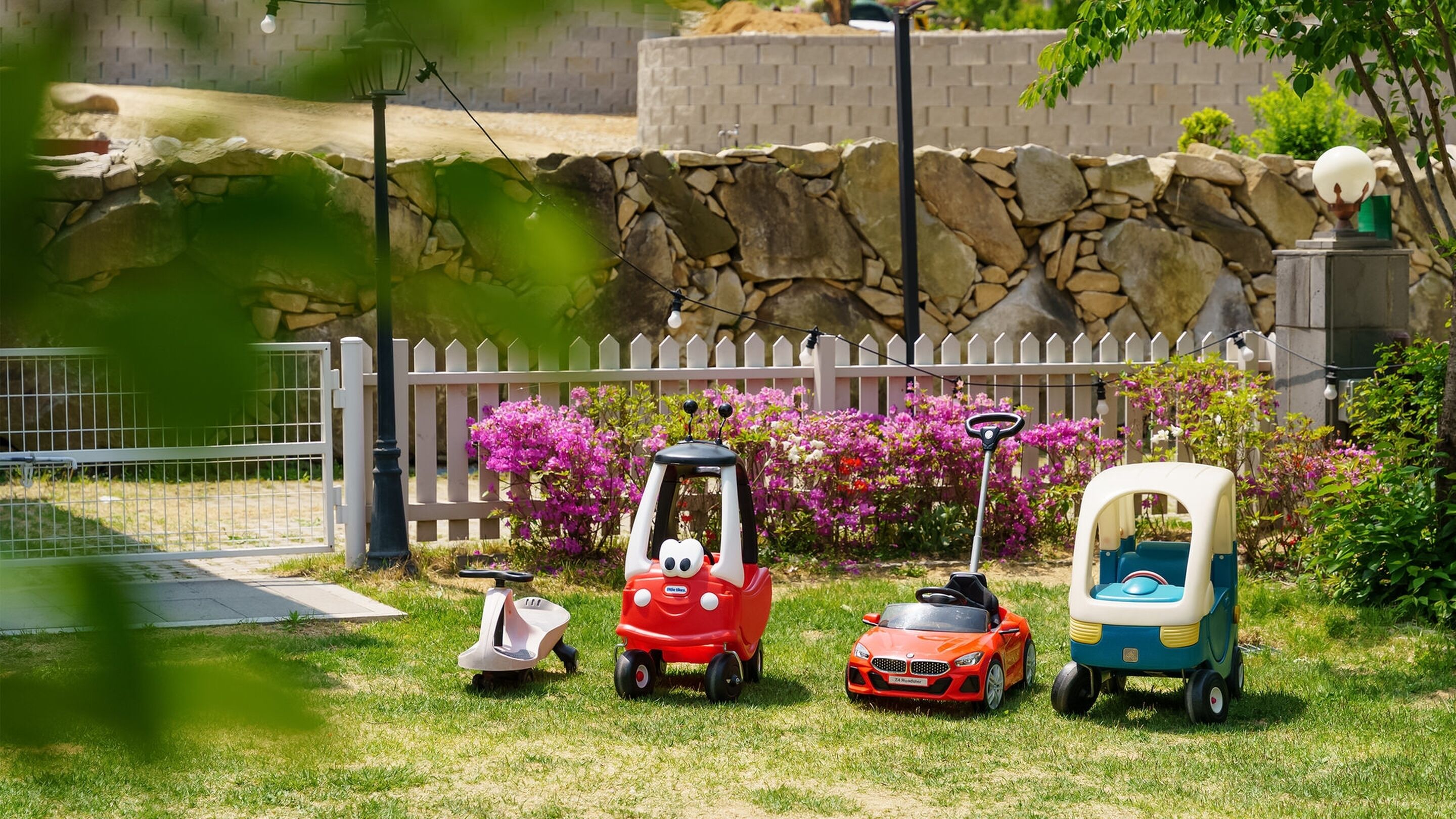 Een tuin met een witte schutting, een stenen muur en diverse speelgoedauto's en een grasmaaier.