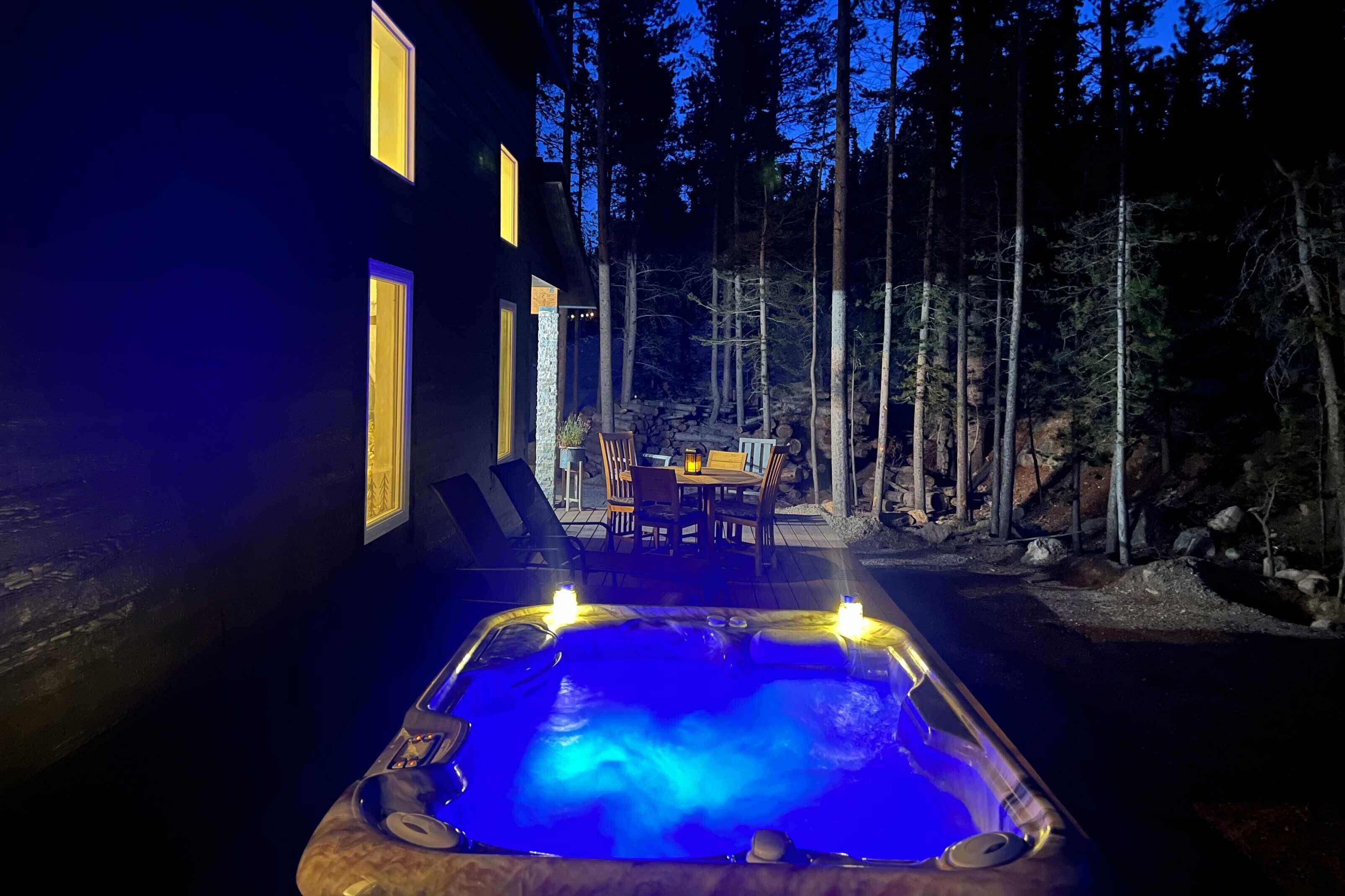 A hot tub in a forested area with a dining table and chairs, surrounded by tall trees.