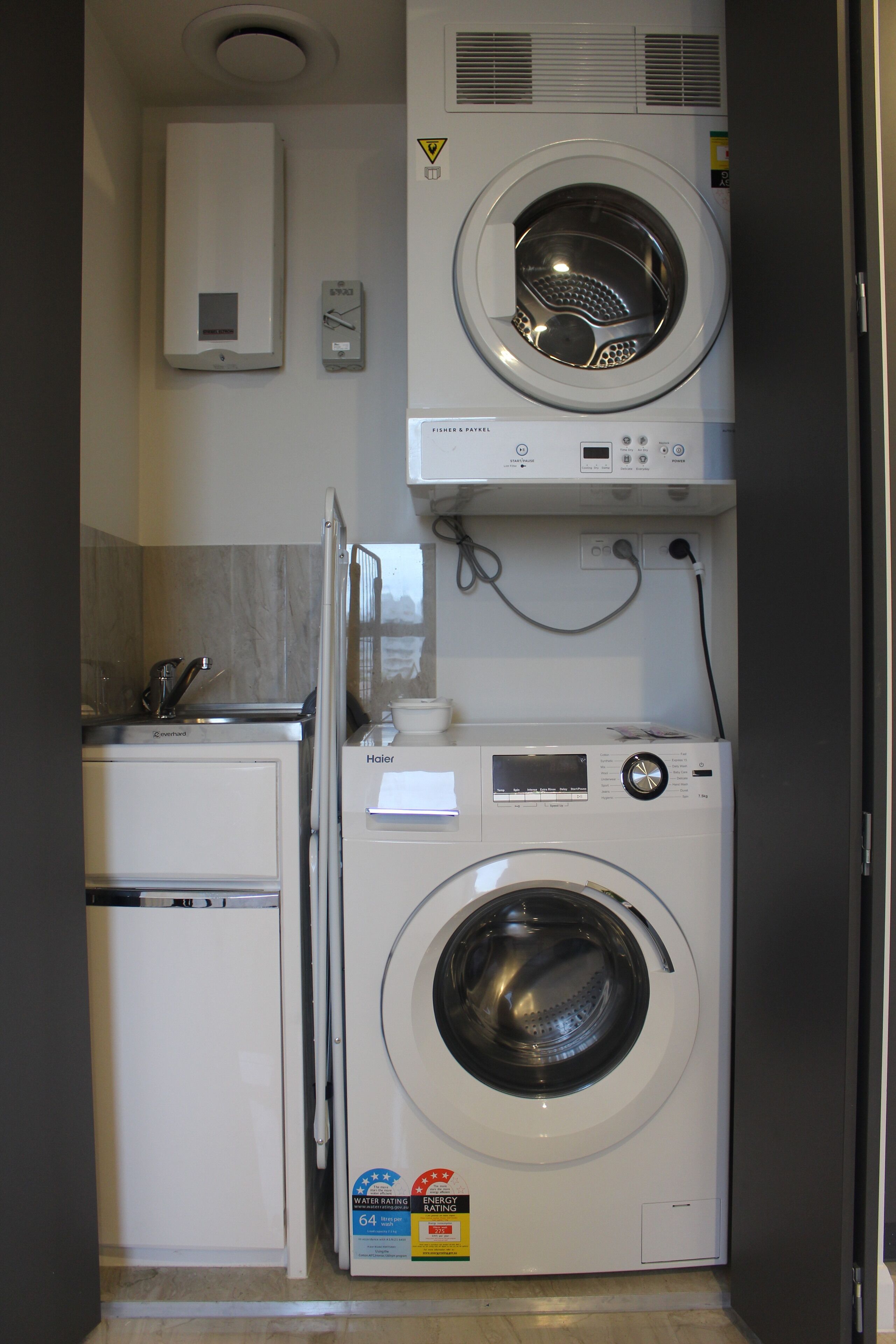 Un área de lavandería compacta con lavadora y secadora apiladas, un refrigerador pequeño y un calentador de agua.