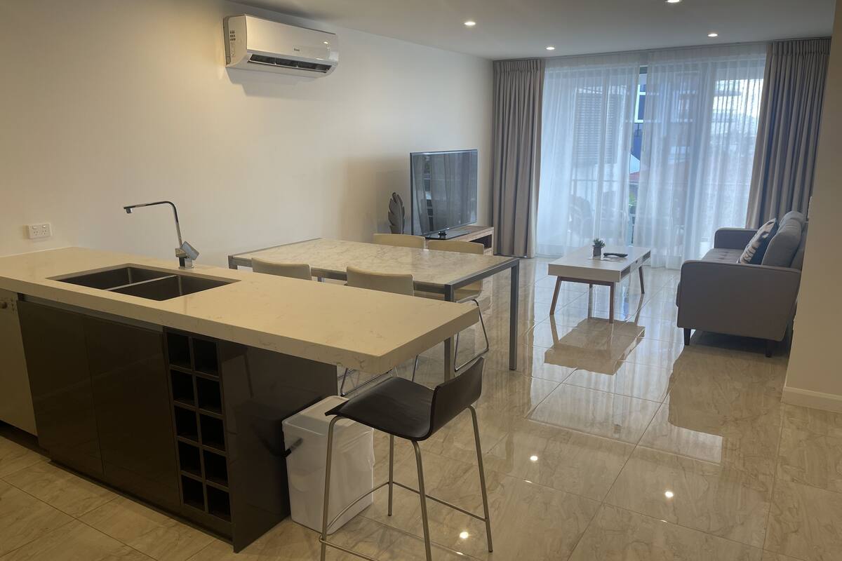 A modern kitchen with a built-in island, a dining area with a table and chairs, a small TV, and a sofa in a living area with a large window.