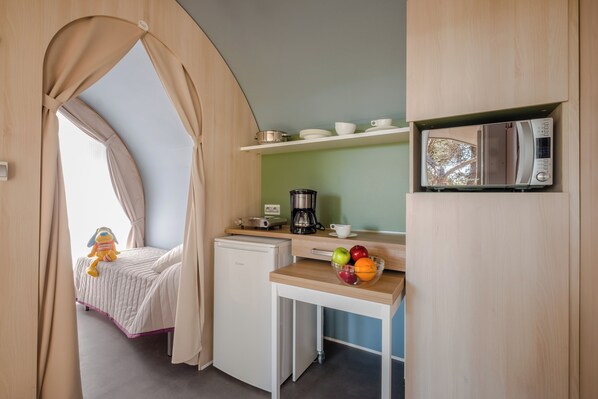 Una kitchenette compacta con microondas, cafetera y un refrigerador pequeño. Se ve una cama con dosel y un peluche al fondo.