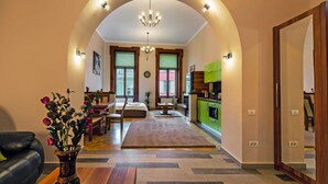 A spacious living area with a sofa, a coffee table, and a vase of flowers. There is a dining table with chairs and a chandelier hanging from the ceiling. The kitchen is visible with green cabinets and a countertop. - Select City Center Apartments Braşov (Brasov)