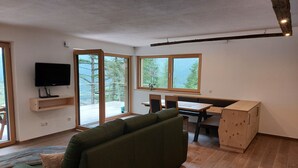 A modern living room with a green sofa, a wooden dining table with chairs, a TV mounted on the wall, and large windows offering a view of greenery outside. - New opening June 2022 - Chalet tree house with private whirlpool (Vandoies)