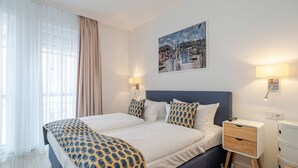 A double bed with patterned bedding, a bedside table, a nightstand, a wall-mounted lamp, and a painting above the bed.