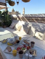 A breakfast setup with a variety of foods and drinks on a table, a sofa with patterned cushions, and a view of a historic building in the background.