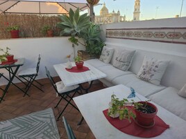 A rooftop terrace with a sofa, two tables, chairs, potted plants, and a view of a historic building.