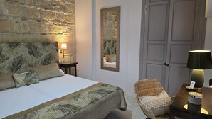 A bedroom with a stone wall, a bed with a leaf-patterned bedspread, a nightstand with a lamp, a wicker chair, and a door. - La Gitanilla Alojamiento & Encanto (Jerez de la Frontera)